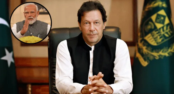 A seated individual in a black and white traditional outfit, with hands clasped, and flags of Pakistan in the background.