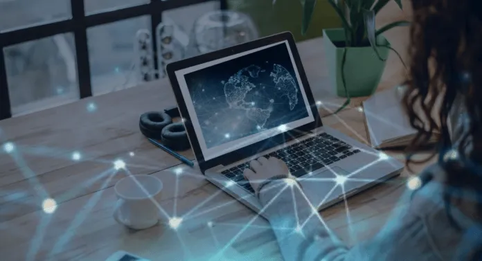 A person typing on a laptop displaying a world map, with a coffee cup and headphones on a wooden table, surrounded by plants.