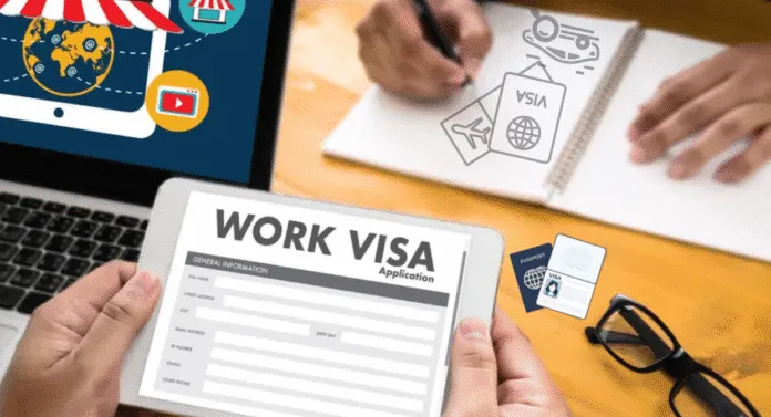 A person holds a work visa application on a tablet, with a laptop and planning materials on a wooden desk.