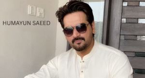 Love Guru Cast Spotlight: Mahira Khan, Humayun Saeed and More 1 A man in a traditional white kurta sits indoors, with modern decor and a large window in the background, showcasing a bright atmosphere.