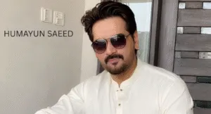 Love Guru Cast Spotlight: Mahira Khan, Humayun Saeed and More 1 A man in a traditional white kurta sits indoors, with modern decor and a large window in the background, showcasing a bright atmosphere.