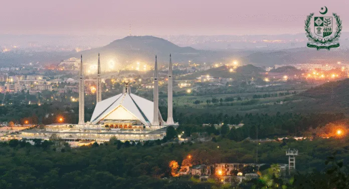 Aerial view of Faisal Mosque in Islamabad, illuminated at dusk, surrounded by lush hills and urban lights, with a logo in the corner.