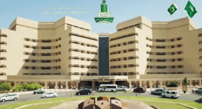 Facade of King Abdulaziz University featuring modern architecture, with cars parked and flags displayed.