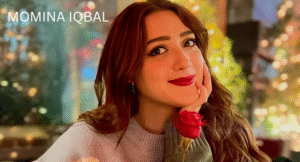 Love Guru Cast Spotlight: Mahira Khan, Humayun Saeed and More 9 Momina Iqbal, with long, wavy hair, sits gracefully, holding a red rose, surrounded by a softly lit, festive background.