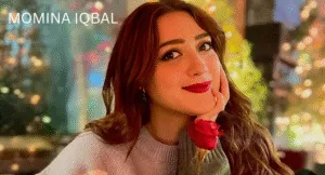 Love Guru Cast Spotlight: Mahira Khan, Humayun Saeed and More 9 Momina Iqbal, with long, wavy hair, sits gracefully, holding a red rose, surrounded by a softly lit, festive background.