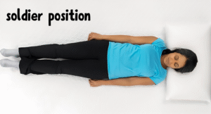 A person lies flat on a bed in the soldier position, wearing a blue shirt and black pants, with a pillow under their head.