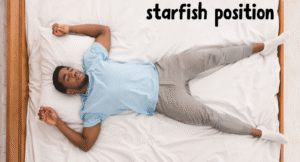 A person lies on a bed in the starfish position, arms and legs spread out, wearing a light blue shirt and gray sweatpants.