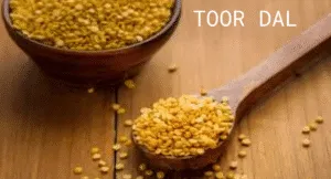 Top Foods That Increase Uric Acid & Effect Your Health: Try Their Alternatives 2 A wooden bowl filled with yellow toor dal (pigeon peas) next to a wooden spoon, scattered on a wooden surface.