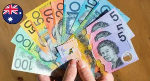 A hand holds a fan of assorted Australian banknotes, including denominations of $5, $10, $20, $50, and $100, against a wooden surface.