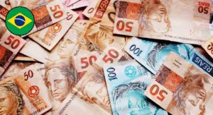 A close-up view of various Brazilian banknotes, featuring denominations of 50 and 100 reais scattered on a surface, with a Brazil flag icon.