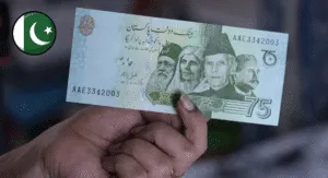 A close-up of a hand holding a 75 Pakistani Rupee note, featuring green and blue hues with text and design elements.