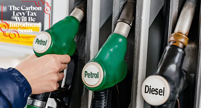 A person holds a green petrol nozzle at a fuel pump, with a diesel nozzle nearby and a sign about an upcoming carbon levy tax.