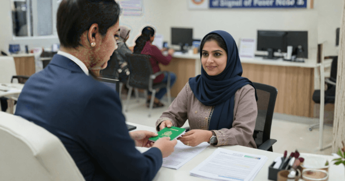 A person hands a green document to a woman in a hijab at a service counter, with computers and clients in the background.